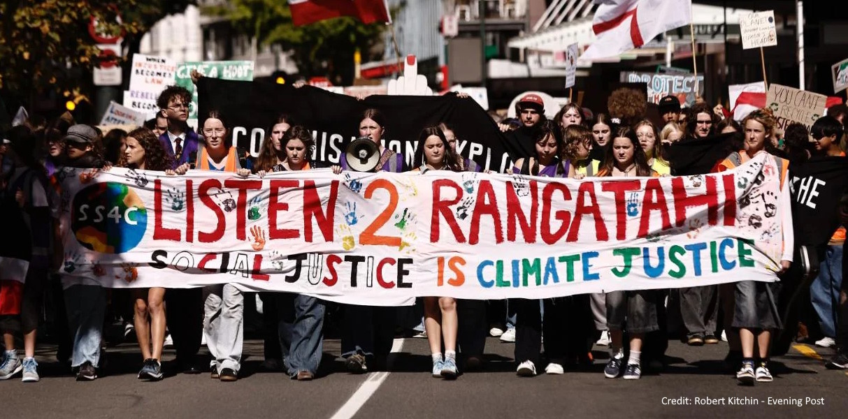 Photo of a protest march coming towards the camera carrying a large banner saying "Listen 2 Rangatahi - Climate Justice".